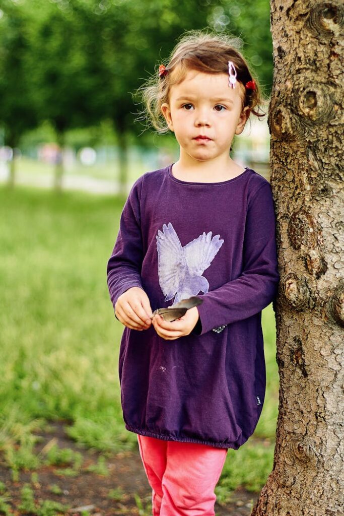 Charming young girl in a purple dress standing by a tree in a park.